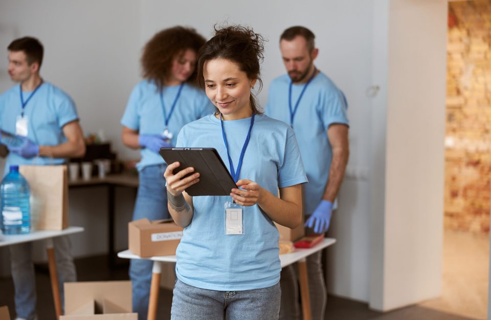 Attractive young female volunteer in blue uniform using tablet pc and smiling while standing indoors. Team sorting, packing items in the background. Charitable foundation concept