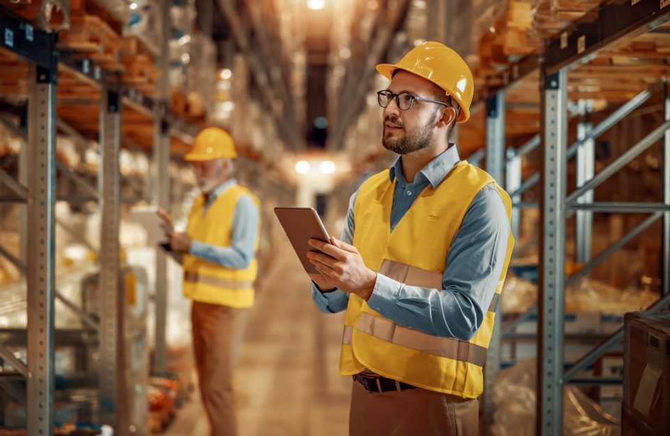 Engineers walking through large warehouse center. They are checking shelves full with boxes and products. Warehouse workers using digital tablet.