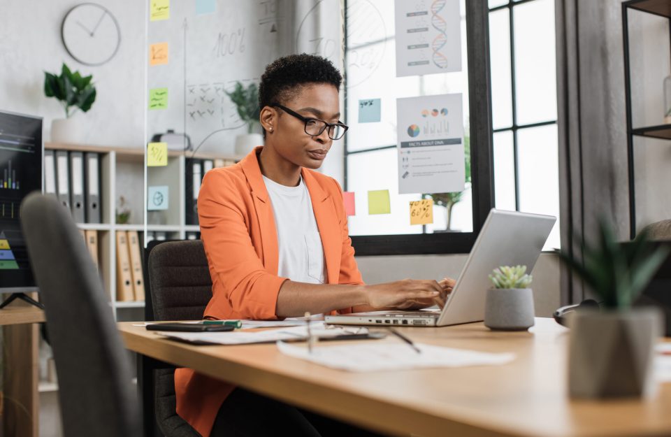 Busy african woman in orange suit sitting at office and using wireless laptop. Big monitor with various charts and graphs on background. Business and finance concept.