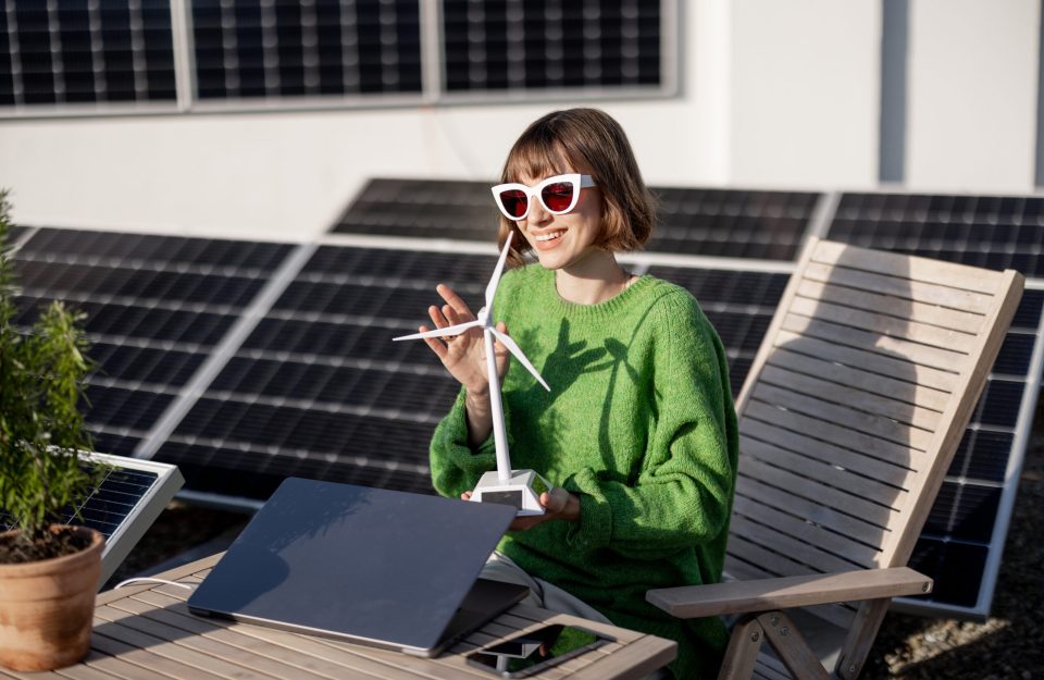 Portrait of a woman with toy wind generator sitting by the table with laptop and flowerpot on the rooftop with solar power plant. Concept of alternative energy and sustainable lifestyle